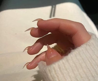 图片特写展示了一只手，这只手的指甲被修剪成异常尖锐且超长的形状，呈现出类似爪子的外观。手部穿着白色毛衣，背景是浅色桌面，隐约可见两根金属餐具。这种夸张的美甲造型可能用于搞笑或视觉冲击效果。