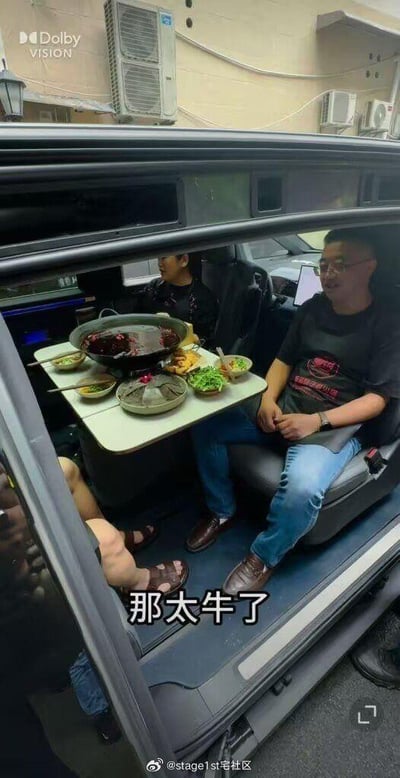 这是一张竖屏的视频截图，画面展示了一辆大空间汽车的内部被临时改造成用餐区，车内摆放着一张小桌，桌上放置着一口正在加热的红油火锅，搭配毛肚、青菜等多种火锅配菜与餐具，几位男士围坐在车内，其中一位戴眼镜的男士坐在汽车座椅上，姿态放松，画面下方配有字幕“那太牛了”，显然是在赞叹这种在车内吃火锅的新奇操作，宽敞的车内空间为这种新奇的用餐场景提供了可能，整体氛围显得十分有趣且充满新鲜感。
