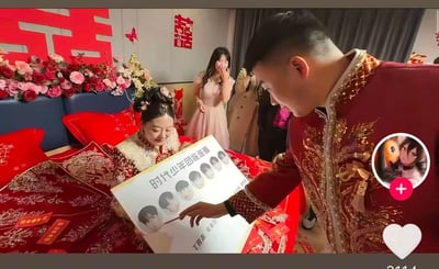 这是一场充满趣味的中式婚礼接亲现场，新娘身着精致秀禾服坐在铺有龙凤刺绣纹样的红色婚床上，手中拿着一块制作好的「时代少年团连连看」游戏板，板上印有丁程鑫、宋亚轩等时代少年团成员的头像及名字。新郎穿着配套的中式礼服，正用手指向游戏板上的头像，似乎在参与这个特别的接亲游戏。背景墙贴着大红双喜字，婚床旁的伴娘看到这一幕掩嘴偷笑，整个场景既有传统婚礼的喜庆氛围，又融入了当下热门的追星元素，反差感拉满，显得格外新颖有趣。
