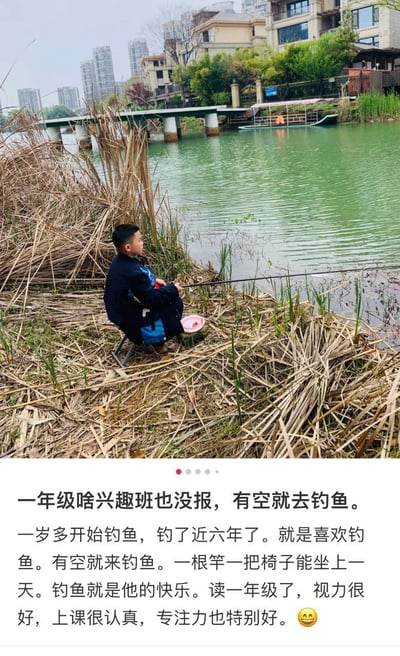 湖边芦苇丛旁，一名一年级小男孩正专注地坐在折叠椅上钓鱼，身旁放着粉色的小鱼桶。背景可见青绿色的湖水、跨湖小桥及远处的居民楼与高楼。配文称男孩从一岁多开始钓鱼，至今近六年，因热爱钓鱼常独自静坐一天，虽未报任何兴趣班，但视力良好且课堂专注力强，展现了无压力的快乐童年。