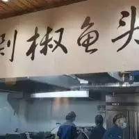 这是一张横版的原始拍摄实景图，画面上方悬挂着一块写有「剁椒鱼头」的书法招牌，字体风格苍劲；下方是餐厅后厨的工作场景，几位身着深色工作服、佩戴帽子和口罩的工作人员正在厨房内忙碌，呈现出餐厅后厨日常运作的真实状态。