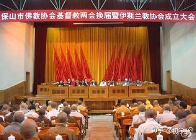 这是一张正式的会议现场原始拍摄照片，画面呈现的是保山市佛教协会、基督教两会换届暨伊斯兰教协会成立大会的现场。前方主席台上就座着会议相关的负责人或代表，台下会场中坐满了参会人员，大家都专注于会议内容。背景是红色幕布搭配多面红旗，顶部悬挂的会议主题横幅清晰点明活动内容，整体场景庄重正式，完整记录了这场宗教协会相关的重要活动。