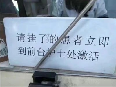 医院场景中的白色指示牌，上面用黑色字体写着“请挂了号的患者立即到前台护士处激活”。指示牌前方有一根金属杆斜向穿过，部分遮挡了文字内容。背景中可见模糊的医院环境和人影，整体呈现真实的医疗场所氛围。