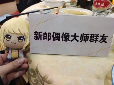 这是国内一场ACGN爱好者婚礼的宴席实拍：餐桌上摆放着写有「新郎偶像大师群友」的专属座位牌，旁边有人手持《偶像大师 闪耀色彩》的角色毛绒玩偶，背景可见五粮液酒瓶与宴席餐具。新郎作为偶像大师爱好者，专门为同好群友设置了席位，将二次元爱好融入三次元婚礼，充满温馨又有趣的氛围。图中无谐音梗或双关暗示。