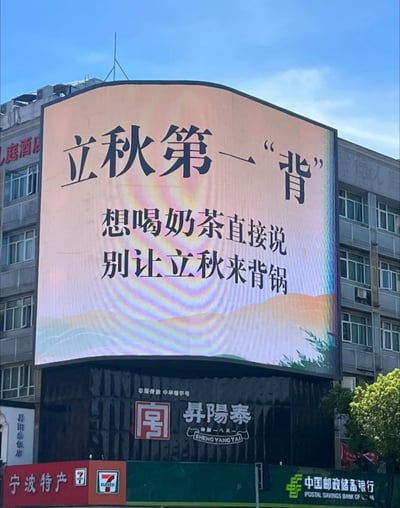 图片拍摄于城市街头的大型LED广告牌，上面用很醒目的字体写着：“立秋第一‘背’”，下面附有“想喝奶茶直接说，别让立秋来背锅”。这句话是网络玩梗，意思是讽刺大家总爱用节气或各种理由掩饰自己想喝奶茶的欲望，调侃了都市年轻人用立秋做借口的无力和幽默。

同时，“背锅”是网络热词，带有双关和谐音梗。既有“背锅”即“被冤枉”之意，也和立秋后贴秋膘（吃东西）的习俗结合，幽默宣传奶茶。