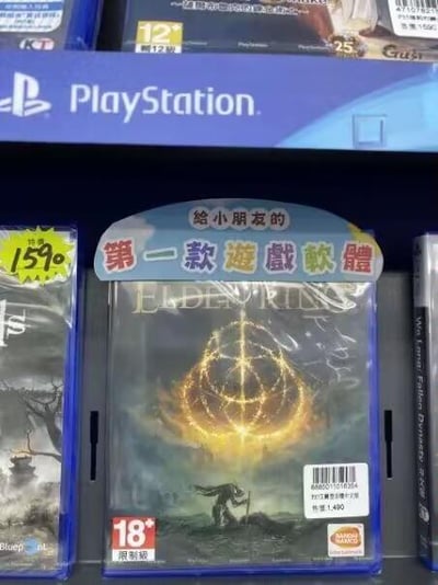 这是一张在游戏实体店拍摄的照片，展示了PlayStation平台的硬核成人向游戏《艾尔登法环》的实体盒。该游戏盒明确标注了18+限制级，却被贴上了“给小朋友的第一款游戏软体”的标签，形成强烈的反差感——《艾尔登法环》以极高的游戏难度和偏黑暗的世界观著称，完全不适合低龄儿童，这个标注制造出戏谑搞笑的效果。