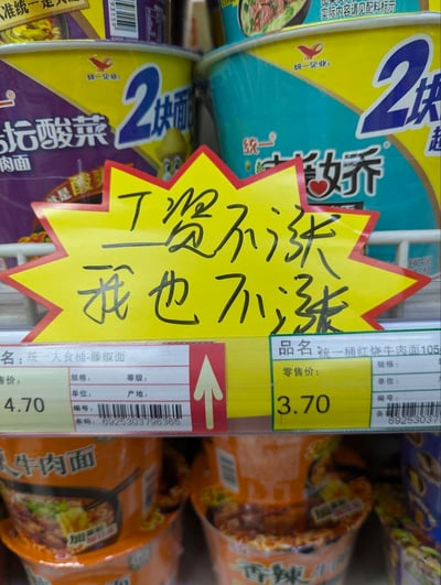这是超市方便面货架的实拍图，货架上陈列着统一企业的多款桶装方便面，包括老坛酸菜牛肉面、藤椒面、红烧牛肉面等产品。货架中间位置贴着一张黄底红边的爆炸形手写标语，上面写着“工资不涨 我也不涨”，下方的价格标签显示统一大食桶藤椒面零售价4.70元，统一桶红烧牛肉面零售价3.70元。标语以幽默接地气的方式，将泡面价格不涨与打工人工资不涨的现状绑定，共情打工人的处境，是直白的吐槽式表达，没有双关或谐音梗。