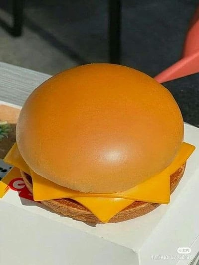 这是一张创意拍摄的静态照片，画面主体是一个模仿经典汉堡造型的物品组合：顶部是一个表面光滑、颜色和汉堡面包高度相近的橙色圆形物体，下方整齐叠放了多片黄色芝士片，最底部是一块类似汉堡底的饼干，整体被放置在一个白色的盒子上。视觉上这个组合酷似一个芝士超多层的汉堡，但实际上是将不同物品创意搭配而成，营造出逼真又有趣的视觉效果，图中没有双关、谐音梗或暗示类内容。