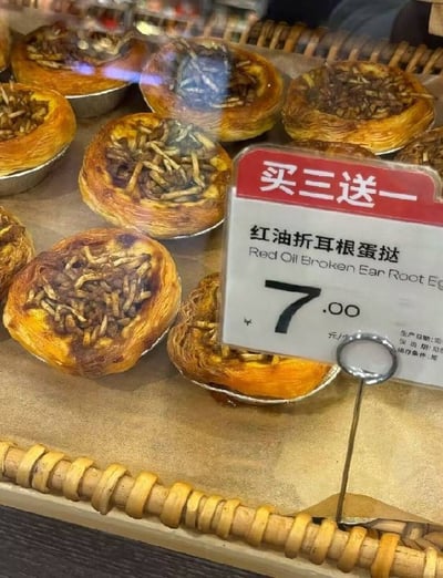 这张图片实拍了线下烘焙展示柜中的猎奇创意新品红油折耳根蛋挞，多个蛋挞放在铝箔托中陈列，每个蛋挞的表面都铺撒了折耳根丝，旁边立着价格标签，标签顶部红色区域标注促销活动为“买三送一”，下方标明产品名称是红油折耳根蛋挞，单只售价7元，这款产品将中国西南特色食材折耳根和西式甜点蛋挞结合，属于比较新奇猎奇的创意美食。