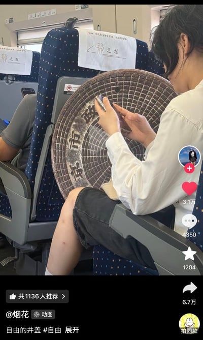 这是一张短视频平台的内容截图，拍摄场景为高铁车厢内：一名穿着白色衬衫、黑色短裤的乘客坐在座位上，腿上放着一个外观高度还原北京市政井盖的圆形背包，包面上清晰印有“北京市政”字样，乘客正低头操作手机。前方座椅靠背上带有高铁“静之旅”的头枕巾，截图右侧是短视频平台的互动按钮，分别显示点赞3.1万、评论4350、收藏1204、转发6.7万，截图底部标注了发布者昵称“烟花”，并配有文案“自由的井盖 #自由 展开”，还有1136人推荐的标识。