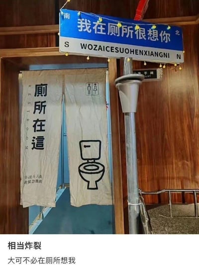 这是一处创意厕所的入口场景：上方模仿网红路牌样式制作了蓝底白字标识，写着“我在厕所很想你”，还标注了南北方向（S、N）和对应拼音；厕所门挂着米白色布帘，左侧布帘印有繁体“厕所在這”、英文“REST ROOM”，还有小字“非战斗人员 请紧急撤离”，右侧布帘画着马桶图案与男女卫生间标识，门旁还有“洗手间”的小标牌；图片下方配有两行文字“相当炸裂”“大可不必在厕所想我”。整体将原本用于浪漫场景的“我在XX很想你”句式，错位放在厕所这个日常场景中，形成强烈反差，制造出搞笑戏谑的效果，没有双关或谐音梗，纯靠场景错位带来幽默。