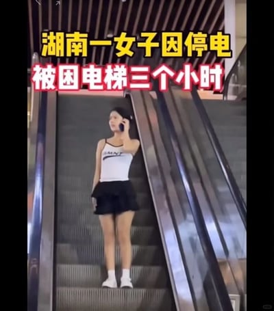 这是一张带有搞笑乌龙性质的图片，画面上方用黄色和红色醒目字体标注“湖南一女子因停电被困电梯三个小时”，但实际图中女子站在停止运行的自动扶梯上，她身着白色吊带上衣、黑色短裙，搭配白袜白鞋，正手持手机打电话，表情较为平静。这张图通过混淆“电梯”和“自动扶梯”的概念制造反差笑点：电梯停电被困是封闭空间的紧急情况，而停止的自动扶梯可正常通行，配文和画面的矛盾形成了趣味调侃效果，属于网络上常见的整活类创作内容。