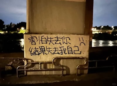 这是一张夜晚户外的实拍图，画面主体是桥体下方的水泥柱，柱面上用黑色颜料手写着“害怕失去你 😢 结果失去我自己”的文字，旁边还搭配了一个难过的表情符号，文字字体潦草，传递出强烈的失意与自我迷失的情绪。背景是泛着灯光倒影的昏暗水面，水泥柱下方有金属护栏，整体环境氛围低落，图片右下角带有小红书平台的水印。