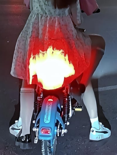 夜晚街头，两名乘客共骑电动车，后方乘客的浅色纱裙垂在电动车尾灯上方，红色的尾灯灯光透过纱裙，呈现出如同裙子底部燃起明亮火焰的视觉效果，形成了逼真又有趣的视觉错位，第一眼很容易让人误以为真的发生了火情。