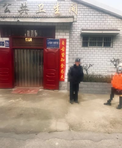 这是一张户外实拍照片，主体是一间砖砌的公共卫生间建筑：门头上方有金属质感的“公共卫生间”五个大字，卫生间红门双开敞开，入口处设有蓝色告示牌标注“公共厕所 免费开放”，门旁墙面贴着一块红底黄字的竖长标语，内容为“自古皆知食为天”，这句话放置在公共厕所旁形成了谐音搞笑的反差效果；入口水泥地面带有无障碍标识，门口铺有写着“出入平安”的地垫，画面右侧有两名站立的路人，都被做了马赛克模糊处理。