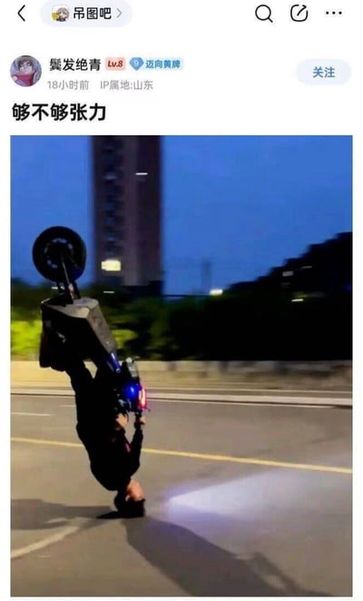 社交媒体平台'吊图吧'用户发布的一张摩托车特技照片。画面中一名男子在黄昏时分的城市道路上，以单手倒立的姿势操控摩托车，摩托车前轮高高翘起，车身倾斜，背景可见高楼与路灯，整体展现出惊险刺激的视觉效果。配文'够不够张力'直接点出内容旨在突出动作的冲击力与表现力。