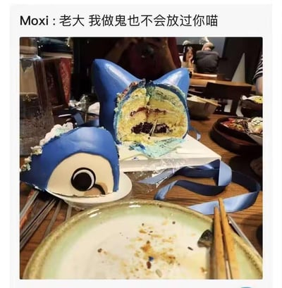 这是一张餐厅餐桌场景的实拍图，定制的卡通角色Moxi造型蛋糕被切分成头部与身体两部分，身体部分被切开露出内部的奶油和夹心馅料，头部单独放置在小盘子中。图上方配文「Moxi :老大 我做鬼也不会放过你喵」，将被切开的蛋糕拟人化，赋予其“被断头”的怨种设定，同时用“喵”字保留角色的软萌感，形成反差感制造搞笑效果。