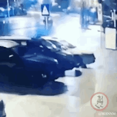 这是一段记录夜晚街道路口事故的动态图，画面中一辆黑色轿车车头严重损毁，停在路口区域，地面因灯光照射有明显反光，背景能看到模糊的行人和道路标识，整个场景呈现出交通事故发生后的混乱状态，氛围惊险。