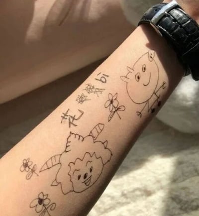 这是一张原始拍摄的静态单图，画面展示了人的小臂区域，上面是用黑色线条绘制的模仿纹身的涂鸦，属于线稿类涂鸦作品。涂鸦包含《喜羊羊与灰太狼》中的两个动画角色：下方是有着标志性卷毛和羊角的懒羊羊，它面带笑意，神态开心；上方是沸羊羊的简化线稿形象，同样面带微笑。涂鸦周围还点缀了几朵简单的小花图案，同时配有手写文字“花臂”和“沸羊bi”，这里运用了谐音梗，将“沸羊羊”的名字与“花臂”的“臂（bì）”发音结合，调侃手臂上的涂鸦为“花臂沸羊羊”，制造出轻松有趣的效果，是一种自娱自乐的趣味创作。