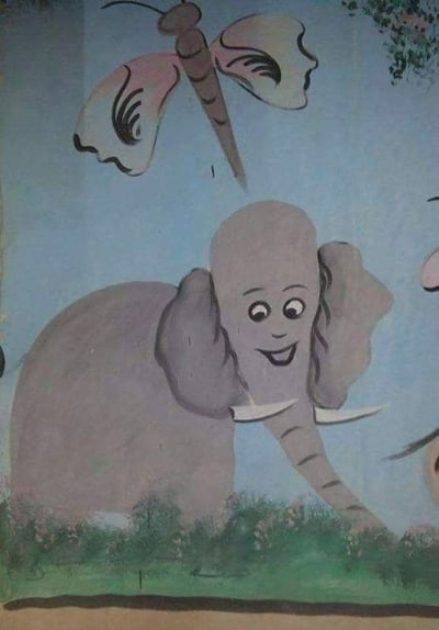 这是一幅童趣风格的手绘卡通画，画面背景是浅蓝色的天空，底部是带有细碎小花的绿色草地。画面主体是一头灰色大象，它的脸部被替换成了人类的脸庞，带着弯弯的眉眼和上扬的嘴角，露出开心的笑容，大象的象牙、长鼻子和大耳朵都保留着原本的形态，长鼻子垂在草地上。画面上方还有一只体型较大的蜻蜓，翅膀是粉白底色搭配黑色纹路，整体画风质朴可爱，没有双关、谐音梗或暗示内容。