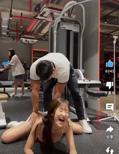 这是一张短视频平台的截图，画面展示了健身房内的趣味日常场景：一位女生趴在地面上做拉伸动作，表情痛苦却又开怀大笑，身后的男生双手按在她的背上，辅助她完成拉伸。背景中还能看到其他正在使用健身器械的人和健身房的专业设备，整体氛围轻松欢乐，充满了生活化的搞笑感。