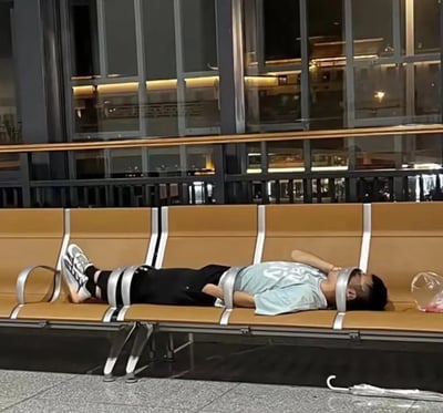 夜晚的公共场所（疑似车站或机场休息室）内，一名男子横躺在一排棕色连排长椅上睡觉。长椅的银色金属扶手恰好从他的腿部、腰部和头部穿过，从特定角度看仿佛将其‘捆绑’在长椅上，形成极具喜感的视觉效果。男子穿着浅蓝色T恤和黑色裤子，脚上是白色运动鞋，身旁地板上放着一个透明塑料袋和一把折叠伞。背景可见大面积玻璃窗，反射出室内的灯光和桌椅陈设。