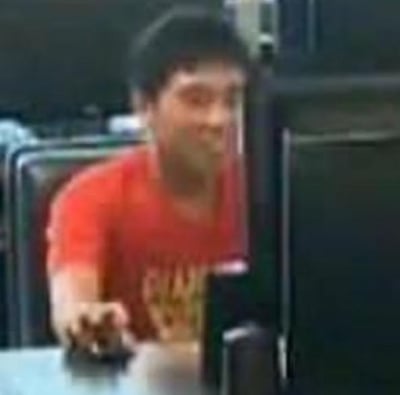 图片中显示一名身穿红色T恤的男子正坐在电脑前，似乎在专注地操作电脑。背景中可以看到多个电脑显示器，暗示这可能是一个办公室、网吧或电脑实验室环境。男子身体微微前倾，右手放在鼠标或键盘位置，表现出正在工作或使用电脑的状态。