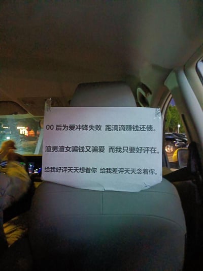 这是一张在滴滴网约车后排拍摄的实拍照片，前排座椅靠背贴着一张打印的白色标语。标语以自嘲的口吻写道：00后为爱冲锋失败，跑滴滴赚钱还债；吐槽渣男渣女骗钱骗爱，而自己只想要乘客的好评；最后用幽默的话术调侃，给好评就天天想着乘客，给差评就天天念着乘客。整体内容结合了网络热梗，风格搞笑又带着一丝无奈，用接地气的方式拉近和乘客的距离，核心目的是获取乘客的好评。