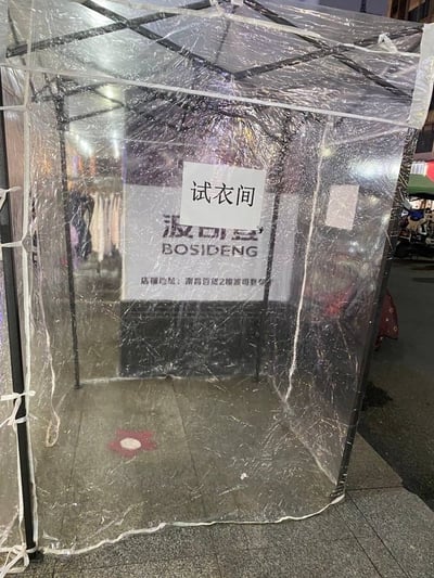 街头出现一个简易试衣间，由黑色金属框架和透明塑料布搭建而成，顶部覆盖着透明塑料。帐篷内可以看到"试衣间"字样的白色标牌，背景中隐约可见"波司登 BOSIDENG"的品牌标识。地面铺有简单的地板，整体给人一种临时搭建、经济实用的感觉，展现了街头商家灵活的经营方式。