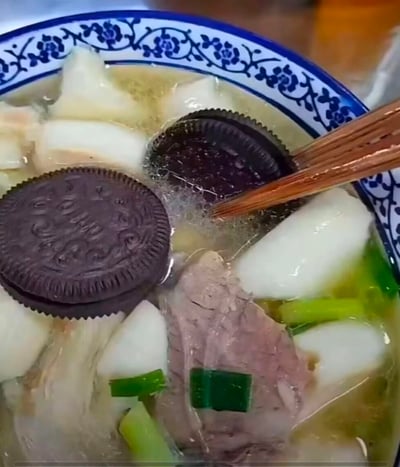 一碗具有传统青花瓷花纹的汤碗中，盛着看似家常的中式汤品，内含肉片、白萝卜块、葱花等食材。然而违和感拉满的是，汤面上漂浮着两块完整的奥利奥饼干，巧克力饼干的深褐色与清亮的汤头形成鲜明对比，筷子正夹起其中一块饼干，仿佛要品尝这奇特混搭。这张图通过将西式甜点饼干强行融入中式传统汤品，制造出荒诞又搞笑的“黑暗料理”视觉效果，引发观众对奇葩食物组合的调侃。