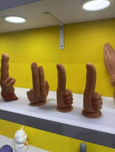 图片展示了一个室内场景，黄色背景墙前的白色架子上摆放着多个棕色的手势造型摆件。这些摆件模拟了不同的手部姿势，包括疑似竖中指、比耶（V字手势）、竖大拇指等。架子左下角还有一个小型的白色兔子造型玩偶。天花板上有两个圆形照明灯，照亮了整个展示区域，整体环境明亮整洁，看起来像是商店橱窗、创意工作室或展览中的展示场景。