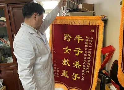 一位穿着白色医生服的男性正在悬挂一面红色锦旗。锦旗上有金色文字，内容为："赠：上海四一医院烧伤整形外科 赵烨博士 及全体医护人员 牛子杀手 跨女救星 二零二四年四月 小斌 敬赠"。背景中可以看到木质橱柜和医疗用品，显示这是一个医院办公室环境。