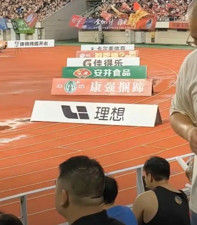 这是一张在体育场观众席拍摄的现场照片，前景是几位正在观赛的观众，中景是橙色的田径跑道，跑道边由近到远依次摆放了多个品牌的广告牌，分别为理想汽车、康强捆蹄、安井食品、佳得乐、卡尔美体育、康师傅喝开水，广告的排列位置刚好让最前方的“理想”品牌名和后方的“捆蹄”二字在视觉上挨在一起，形成谐音“理想亏提”的趣味效果，用来调侃理想汽车降价后刚提车的车主出现利益损失的相关事件，背景是坐满观众的体育场看台，还悬挂有其他赛事相关的宣传横幅。