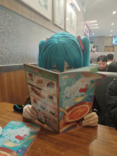 在一家连锁餐厅内，一位爱好者cos成虚拟歌手初音未来，戴着初音的头套与手套，双手举着餐厅的菜单挡在脸前，仅露出眼睛，仿佛在认真查看菜单又带着一丝腼腆。背景可见其他用餐的顾客，餐厅是现代简约的装修风格，菜单上印有餐品图片、介绍以及搭配餐品图案的世界地图设计。整体画面将二次元cos与三次元日常场景结合，氛围轻松有趣，充满可爱感。图中无谐音梗或双关内容。