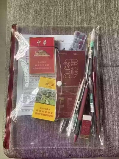 这是一个用于考试的透明文件袋，里面配备了晨光品牌的全套考试文具，包括中性笔、涂卡笔、橡皮、尺子，还有文化考试的准考证及考试时间表，除此之外还额外放入了中华、南京两个品牌的香烟，形成备考用品与香烟的有趣组合，没有谐音梗或双关含义，只是考生的个性化装备搭配。