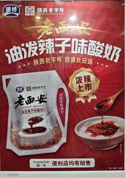 这是一张竖版的银桥品牌新品宣传海报实拍图，整体采用喜庆的大红色作为主背景，顶部标注银桥品牌及“陕西老字号”标识，核心宣传产品为老西安油泼辣子味酸奶，打出“陕西老字号 地道长安味”的宣传语。海报左侧展示了净含量180g的该款酸奶产品实物包装，包装上印有西安大雁塔、祥云等地域特色元素。海报右侧配有金色的“泼辣上市”徽章标识，同时展示了木勺将带芝麻的油泼辣子浇在白酸奶上的产品效果示意图。海报底部标注该产品可在“每一天”连锁便利店购买，右下角小字标注宣传语中的老字号所指为银桥品牌，左下角带有分众电梯海报的标识，属于将地方特色风味与常规乳制品结合的创新猎奇产品宣传物料。