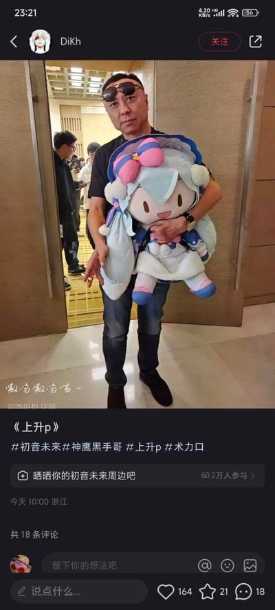 社交媒体截图中，一名戴墨镜、穿黑色T恤和牛仔裤的男子（神鹰黑哥）双手抱着一个超大号初音未来毛绒玩偶。玩偶有着浅蓝色长发、粉色蝴蝶结发饰和白色绒球，造型可爱。背景为室内活动场所，可见其他人员走动。帖子配文“嗷呜嗷呜~”，标题《上升p》，并带有多个相关话题标签。男子严肃的表情与怀中可爱的动漫玩偶形成鲜明反差萌。