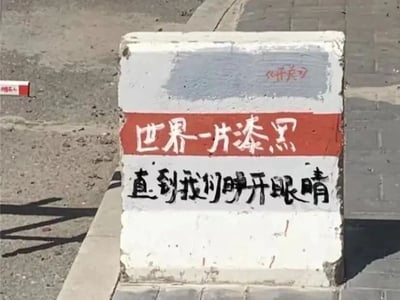 这是一张原始拍摄的静态图，画面展示了一个放置在城市人行道与路面交界处的水泥隔离墩，墩身表面有明显的磨损和污渍，显得有些陈旧。有人在墩身进行了手写涂鸦创作：顶部右上方用红色笔迹标注了《开关》，中间区域以红底白字呈现“世界一片漆黑”，下方则是黑字书写的“直到我们睁开眼睛”。这段文字出自歌曲《开关》的歌词，传递出一种从混沌迷茫中觉醒、迎来光明与希望的积极治愈情绪，将原本普通的城市公共设施，变成了承载文艺与温暖的街头小景。