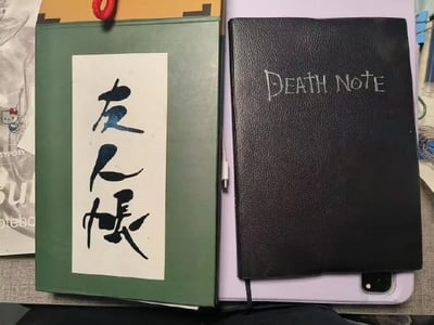 图片展示了两本具有动漫代表性的笔记本并排摆放的场景。左侧为绿色封面笔记本，封面上贴有白色标签，手写蓝色毛笔字“友人帐”，源自日本动漫《夏目友人帐》，整体风格清新温暖；右侧为黑色皮质笔记本，封面用白色字体写着“DEATH NOTE”，源自经典动漫《死亡笔记》，风格神秘暗黑。背景中可见Hello Kitty图案的纸巾、紫色笔记本边缘及部分电子设备，营造出日常桌面的真实感。两本笔记本形成鲜明对比，分别代表了治愈系与暗黑系的动漫气质。