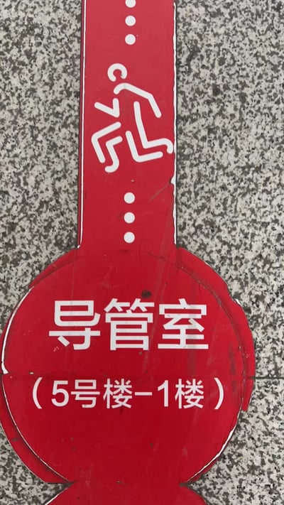 一张医院内的红色指示牌实拍照片，背景为灰色花岗岩墙面。指示牌主体呈竖长形，顶部有白色奔跑人形图标，下方圆形区域内标注白色文字“导管室（5号楼-1楼）”，用于指引前往5号楼1楼导管室的位置。