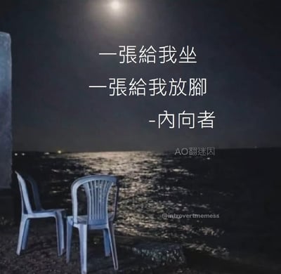这张图以夜晚海边为背景，月光照射在海面形成粼粼波光，岸边砂石地上摆放着两把白色塑料靠背椅，图中配文以调侃的口吻描述内向者的社交习惯：一张椅子用来自己坐，另一张椅子用来放脚，幽默展现了内向人群不想被陌生人打扰、宁愿占用额外座位也不愿和陌生人邻坐的抗拒社交的心态。