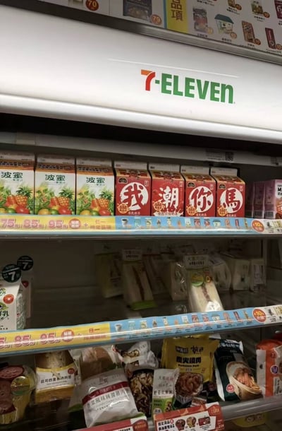 这是一张在7-ELEVEN便利店冷藏货架前拍摄的照片，货架上层左侧摆放着三盒波蜜果菜汁，右侧的四盒饮料包装上的汉字依次为“我”“潮”“你”“馬”，通过谐音“潮=操”“馬=妈”，刚好组成了一句低俗粗口，利用商品摆放的偶然巧合制造出反差搞笑的效果。
这是典型的利用商品文字的偶然排列形成的谐音梗，用日常便利店的普通场景和粗口内容形成强烈反差，带来恶搞趣味。