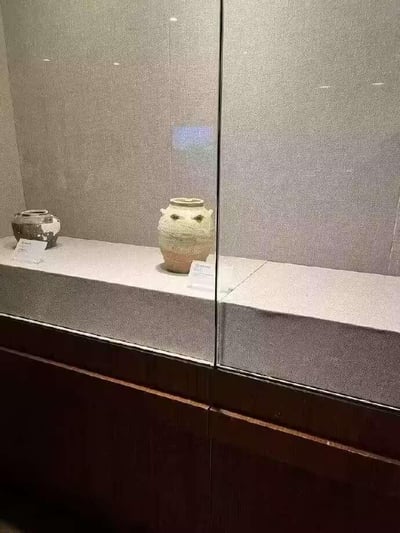 这是一张博物馆展柜的实拍照片，展柜内陈列着两件古代陶罐类文物。其中位于画面中间的陶罐，其肩部的系耳与装饰纹路恰好组合成类似卡通人物眼睛和嘴巴的形态，形成了仿佛带着惊讶表情的视觉效果，让原本古朴严肃的文物意外透出萌感，充满趣味。