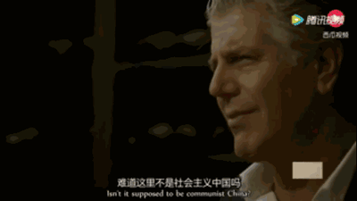 这是一张横屏的影视截图，画面右侧呈现一位白发老年男性的侧脸，他神情略带疑惑与质疑，目光投向画面左侧；画面下方配有中文字幕“难道这里不是社会主义中国吗”，以及对应的英文翻译“Isn't it supposed to be communist China?”；画面右上角带有腾讯视频和云视听极光的平台水印，整体构图清晰，属于带有字幕的真人影视画面截图。