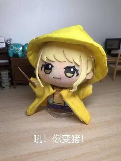 图片中展示了一个可爱的黄色巫师风格玩偶，它有着金色的头发、大大的眼睛和黄色的斗篷与帽子。玩偶手持一根小魔杖，表情略带严肃地站立在木质桌面上。背景中可以看到房间的一角，有蓝色的大象摆件和电脑等物品。图片底部配有文字“吼！你变猪！”，为整个画面增添了趣味性和魔法氛围。
