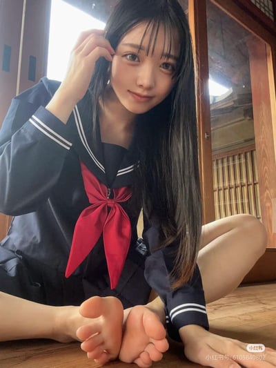 图中是一位身着藏青色水手款JK制服的长发少女，坐在日式木质房间的地板上。她的制服带有白色双条纹装饰的水手领，搭配醒目的红色大领结，左手轻撩额前发丝，双脚自然向前伸展，对着镜头露出温柔甜美的微笑。背景是带有木质格栅的日式门窗，整体画面充满日系校园感，氛围清新治愈。
