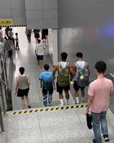 这是在国内地铁站的下行通道内拍摄的画面，画面中走在前方的两位穿白背心背背包的男生，与他们身后的两人因视角巧合形成视觉错位，看起来像是一个拥有四只手臂的壮汉，场景充满趣味性。

这是典型的**视觉错位**效果，利用人物的位置和拍摄视角的巧合，制造出荒诞的视觉假象，让人第一眼产生认知错觉。