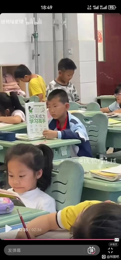 这是一张小学课堂的视频截图，画面中是一间普通的小学教室，多数学生处于放松状态：有的趴在课桌上休息，有的在教室后方活动。而画面中间的小男孩正坐在课桌前，举着一本名为《30天成为学习高手》的书籍，表情茫然略显无奈，和周围同学的松弛状态形成反差，营造出一种滑稽有趣的氛围，仿佛在表达想提升学习能力却又力不从心的情绪。