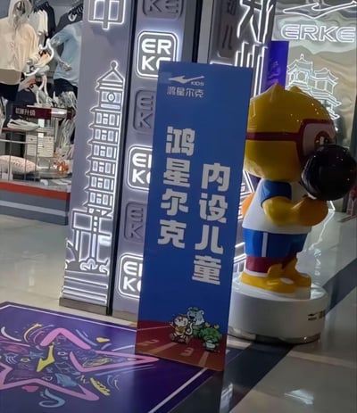 这是一张鸿星尔克线下门店的实拍照片，画面主体是一块蓝色立式引导牌，上面标注着「内设儿童 鸿星尔克」以及「鸿星尔克KIDS」的品牌标识，牌面底部还有品牌卡通形象；引导牌旁立着一个戴着眼镜、抱着篮球的黄色卡通公仔，是鸿星尔克儿童线的IP形象；背景是带有发光效果的鸿星尔克品牌标识和郑州地标二七塔造型的装饰柱，左侧区域能看到门店内的服饰陈列，整体场景用于告知到店顾客该门店设有儿童服饰专区，没有双关、谐音梗等特殊内容。