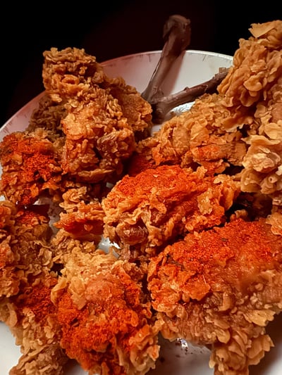 这是一张近距离拍摄的美食实拍图，白色盘子里堆放着多只炸鸡腿，炸鸡外皮呈现出蓬松酥脆的鳞片纹理，部分鸡腿表面撒有鲜亮的红辣椒粉，油润金黄的色泽在深色背景的衬托下格外诱人，看起来外酥里嫩，充满了让人垂涎的食欲感。